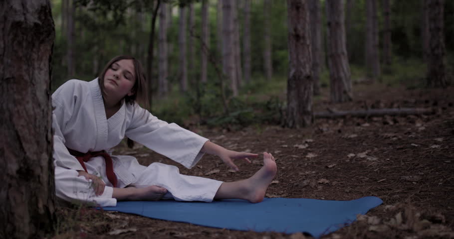 Girl in karate uniform stretches on a yoga mat in a peaceful forest, practicing martial arts discipline.