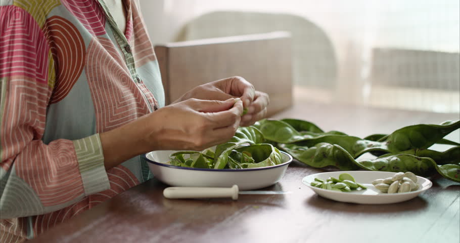 Close up of Middle Aged Asian Woman Hands Peeling Parkia Speciosa Stink Bean with Knife into White Plate on Table,  Faceless Female Preparing Bitter Bean Food Ingredient in Morning Kitchen Room