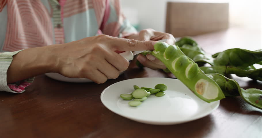 Close up of Middle Aged Asian Woman Hands Peeling Parkia Speciosa Stink Bean with Knife into White Plate on Table,  Faceless Female Preparing Bitter Bean Food Ingredient in Morning Kitchen Room, and regional culinary cultures.