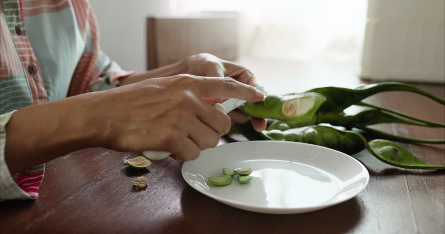 Close up of Middle Aged Asian Woman Hands Peeling Parkia Speciosa Stink Bean with Knife into White Plate on Table,  Faceless Female Preparing Bitter Bean Food Ingredient in Morning Kitchen Room