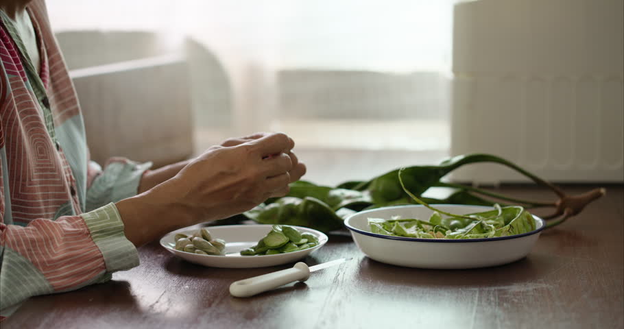 Close up of Middle Aged Asian Woman Hands Peeling Parkia Speciosa Stink Bean with Knife into White Plate on Table,  Faceless Female Preparing Bitter Bean Food Ingredient in Morning Kitchen Room, and regional culinary cultures.