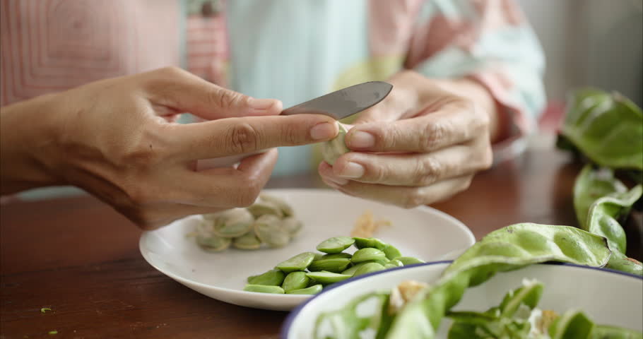 Close up of Middle Aged Asian Woman Hands Peeling Parkia Speciosa Stink Bean with Knife into White Plate on Table,  Faceless Female Preparing Bitter Bean Food Ingredient in Morning Kitchen Room
