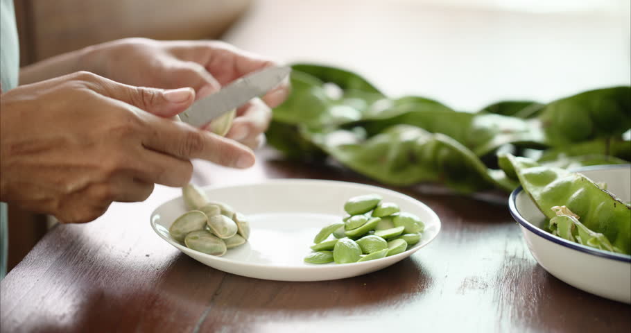 Close up of Middle Aged Asian Woman Hands Peeling Parkia Speciosa Stink Bean with Knife into White Plate on Table,  Faceless Female Preparing Bitter Bean Food Ingredient in Morning Kitchen Room, and regional culinary cultures.