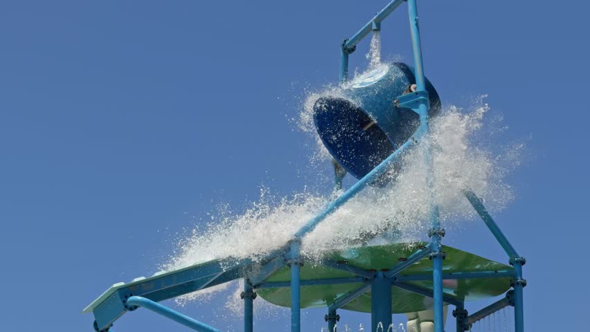 Static medium shot of a giant water bucket tipping over and spilling a massive splash onto a colorful kids play playground structure at a sunny aqua park resort.