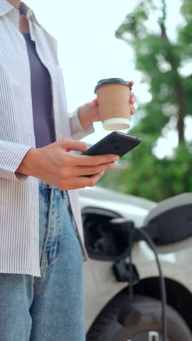 Close-up of happy Asian woman holding coffee and using a smartphone while charging her electric car at home. Concept of modern eco lifestyle green tech and sustainability. Vertical Shot
