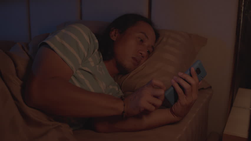 A man uses his smartphone while lying on a bed in a dark room at night, bedtime scrolling, insomnia, and excessive social media use.