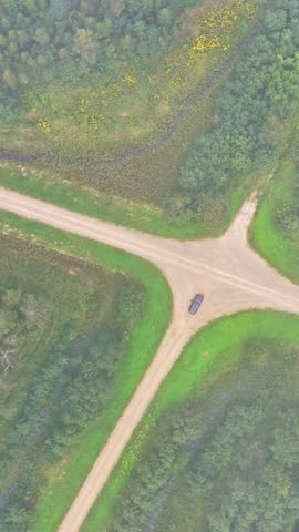 Aerial vertical shot descends above single car parked at rural dirt road crossroad surrounded by green meadow and forest - top down shot