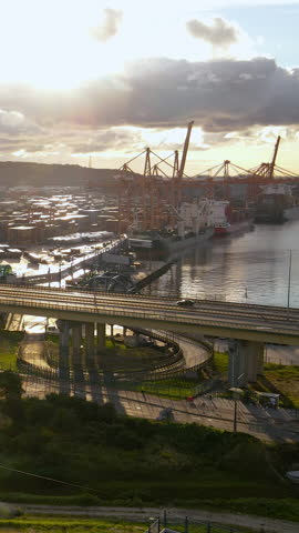 Cinematic vertical shot of a busy marine cargo terminal with cranes, ships, and highway bridge during sunset. Global trade and logistics concept.