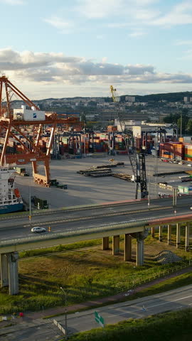 Aerial view of a busy maritime shipping port in Gdynia, Poland, featuring heavy duty cranes, cargo ships, and a highway bridge with traffic during golden hour.
