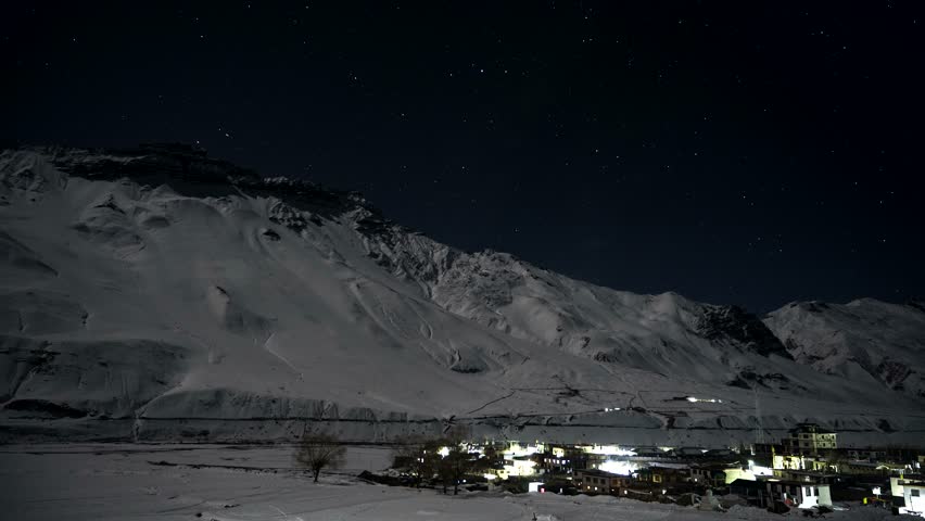 Timelapse footage capturing circular star trails rotating across dark night sky above snow covered mountain range and brightly lit village located in remote himalayan valley landscape.