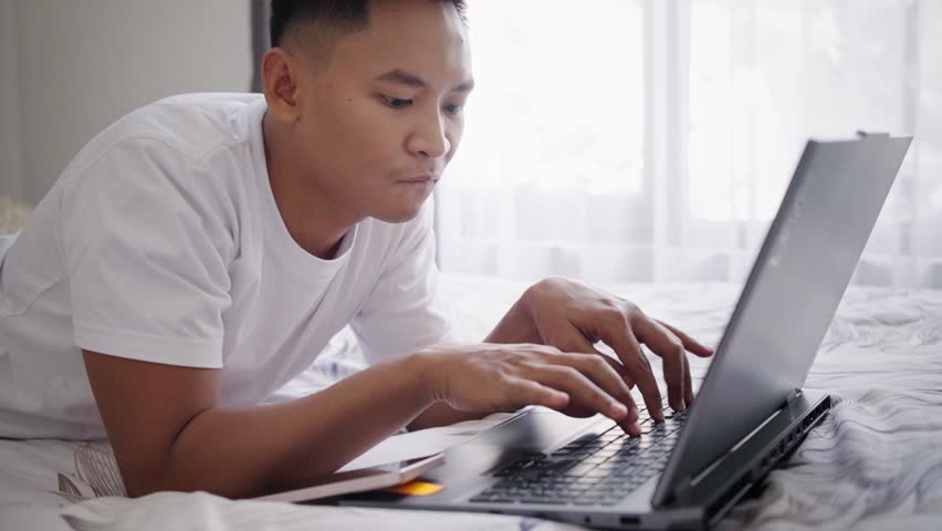 Young Asian Man Working or Studying Remotely on Laptop in Casual Bedroom Workspace Concentrating on Screen