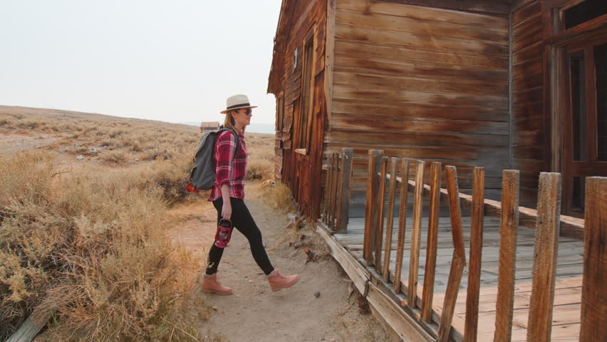 Girl walks through old abandoned ghost town in America exploring weathered wooden buildings. Female traveler in plaid shirt and straw hat discovers historic western architecture with desert landscape.