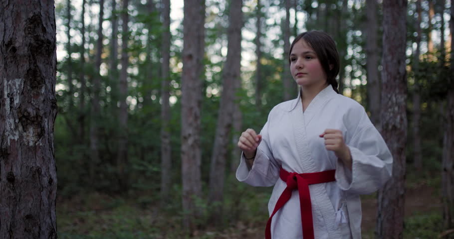 Young girl in a karate gi with a red belt practices martial arts in a serene forest setting.