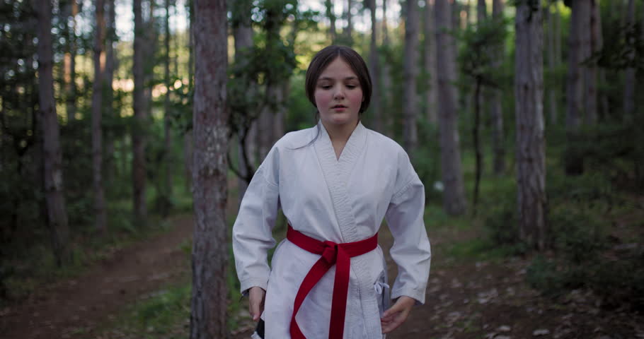 A determined young girl in a karate gi with a red belt practices martial arts in a serene forest.