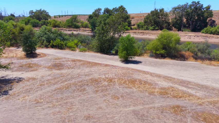 Ascending aerial drone footage rising over the San Joaquin River in Fresno, California, revealing people swimming in the water alongside lush riparian vegetation, trees, and bushes. A dirt hiking trail runs along the riverbank, offering outdoor recreation access, with power lines and transmission towers visible in the distance. This dynamic, cinematic aerial reveal showcases the tranquil river, tree canopy, and scenic Central Valley landscape from a birds eye perspective.