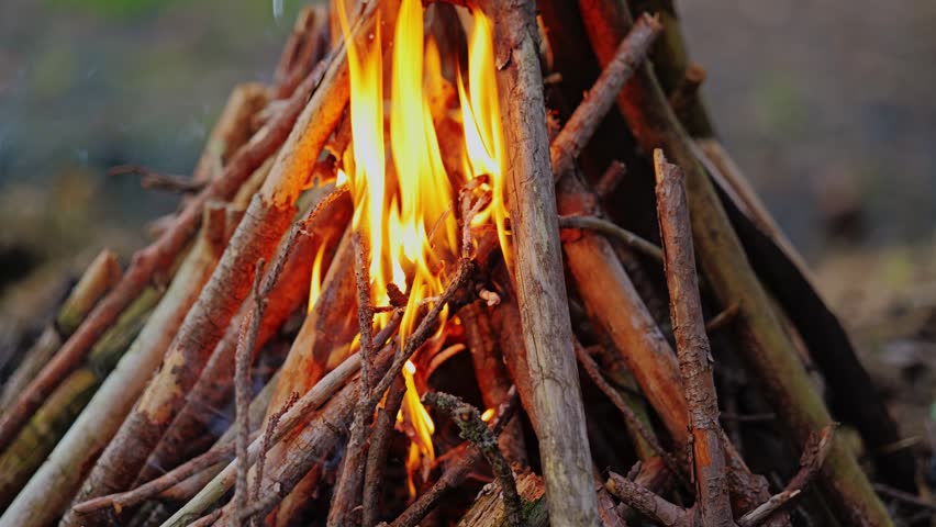 Close up shot shows fire burning in a teepee structure as human hand places thin dry sticks onto glowing embers to maintain warm flames during outdoor adventure in wilderness.