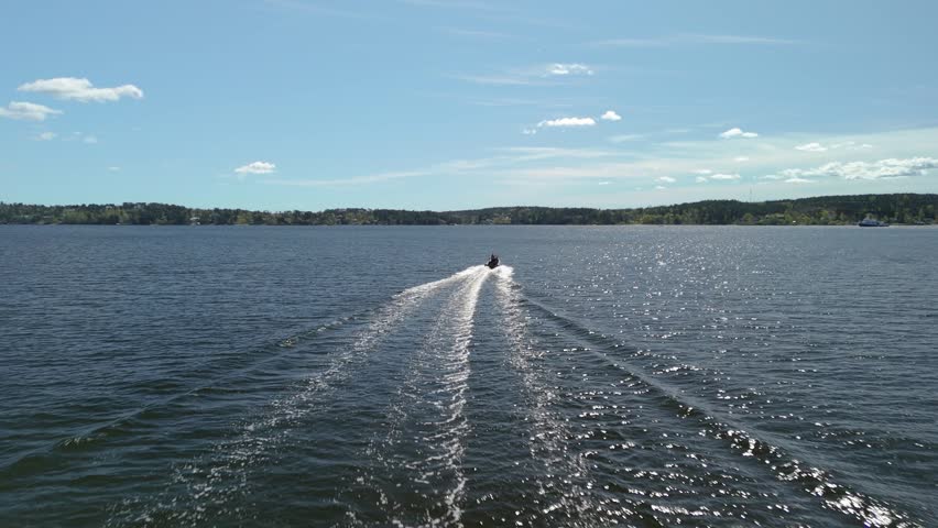 Aerial cinematic footage capturing jet ski rider moving rapidly over deep blue water creating foam wake trail on sunny day against distant coastal forest landscape background travel sports.