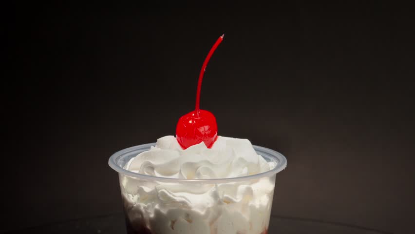 Slow motion close up of delicious chocolate mousse topped with whipped cream and bright red maraschino cherry spinning on rotating platform against dark studio background.