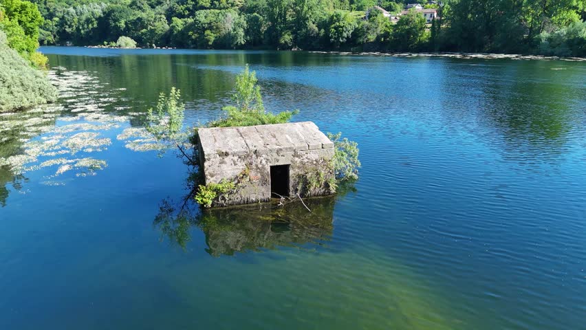 Old stone structure partially submerged in the middle of a river. Calm blue water reflecting the sky and the surrounding green forest