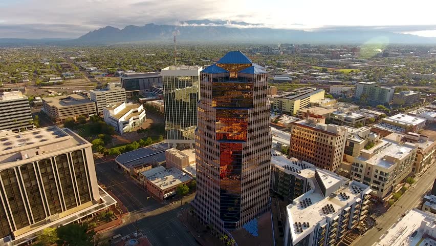 Aerial drone footage capturing cityscape of downtown Tucson Arizona featuring modern glass skyscrapers reflecting morning sun light during sunrise cinematic motion over urban landscape.