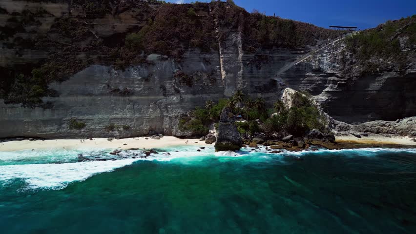 Dynamic aerial view of a secluded beach with dramatic cliffs and crystal clear blue water. Waves crash gently onto sandy shore. Slow motion motion.