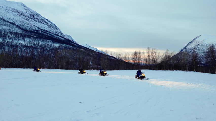 People on snowmobiles with headlights on driving in line across snowy landscape in Tromso, Norway. Aerial drone at low altitude, copy space