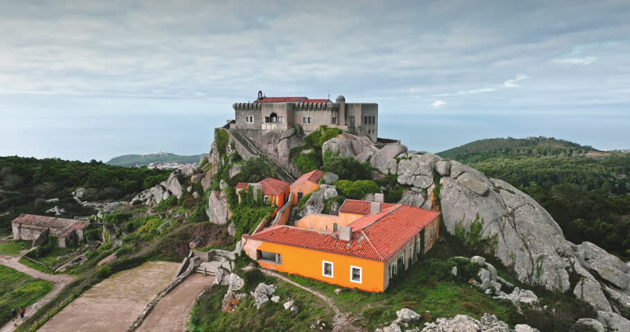Aerial panorama of historic Sanctuary of Peninha complex on rocky peak overlooking the Atlantic Ocean, featuring ancient buildings nestled among rugged landscapes and lush greenery of Sintra, Portugal