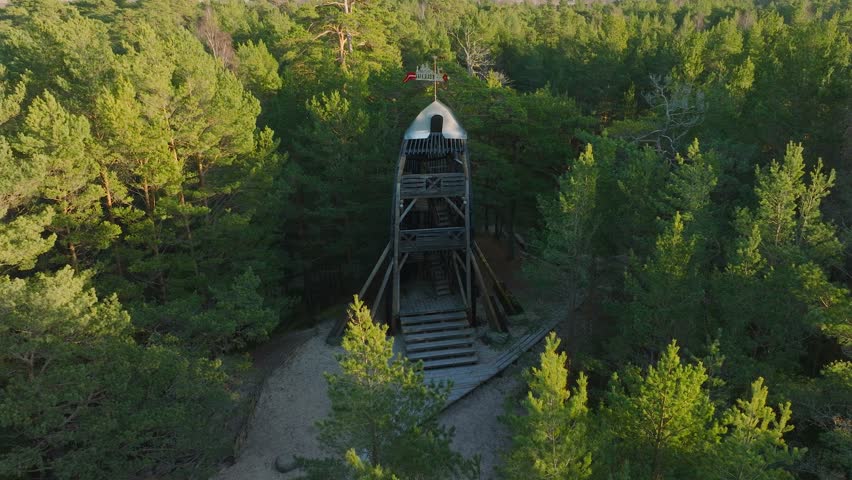 Dynamic aerial shot flying backward from rustic wooden lookout tower nestled within vast green forest. Morning sunlight illuminates treetops in scenic wilderness landscape footage.
