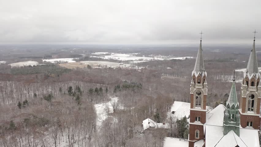 Dramatic aerial flight moving past gothic church towers over snowy forest hills, wide cinematic vista of wintry horizon under gray overcast sky, dynamic religious architecture footage.