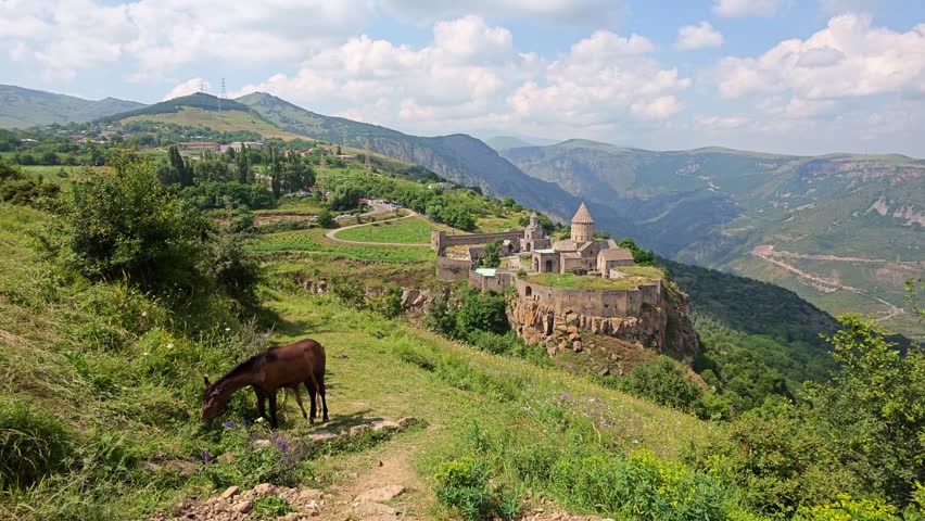 Horse grazes on hillside near ancient monastery ruins. Mountainous terrain stretches into distance