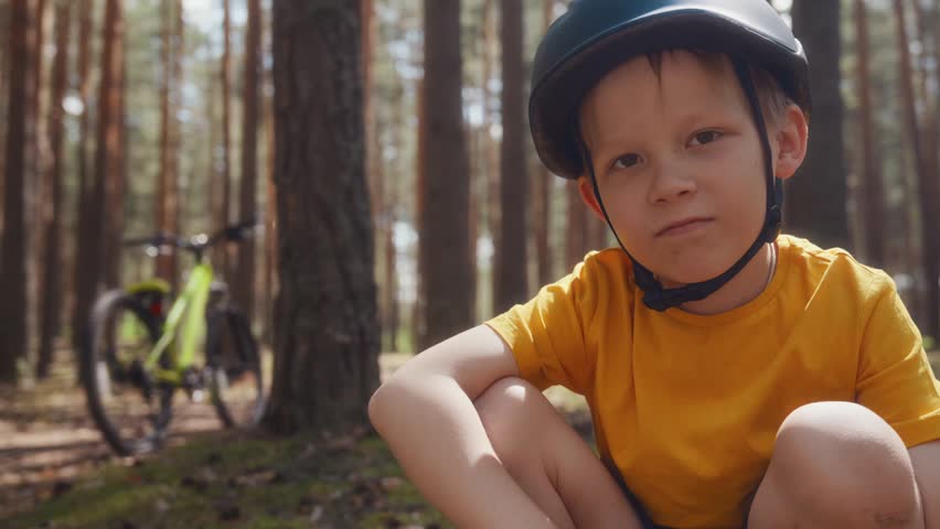 Close portrait of an eight year old boy in a bike helmet showing thumbs up to the camera in a sunny pine forest with his bicycle behind him as a positive call to cycling. High quality 4k footage