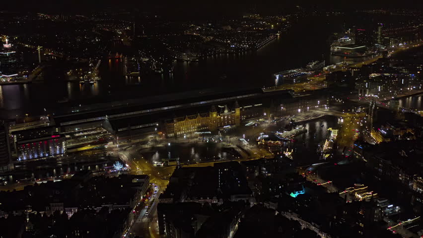 Aerial night view of Amsterdam Central Station, Netherlands, with the illuminated railway terminal, ferry docks on the IJ river, canal bridges, holiday lights and traffic in the city center.