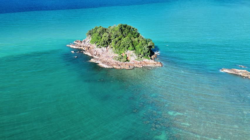Experience the remarkable beauty of Punggur Island in Bangka through stunning aerial drone footage that captures an untouched tropical destination surrounded by brilliant turquoise water and impressive granite coastlines. This cinematic video showcases a small forested island rising above crystal-clear seas, where rugged rock formations meet shallow coral reefs and transparent coastal water. The elevated perspective reveals intricate underwater textures, vibrant marine colors, and the harmonious contrast between dense tropical vegetation and weathered stone. Gentle waves move across the surrounding ocean while sunlight enhances the vivid blue and emerald tones, creating a peaceful atmosphere that reflects the natural charm of this secluded island. Wide panoramic views emphasize the expansive horizon and the pristine environment, making the footage suitable for travel promotions, tourism campaigns, environmental documentaries, destination marketing, luxury vacation advertisements, television productions, and creative storytelling. Every frame highlights the geological uniqueness of Bangka