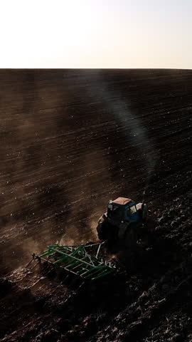 Agricultural tractor plowing dark soil field under clear sky, creating furrows and dust clouds while sunlight casts long shadows across the landscape