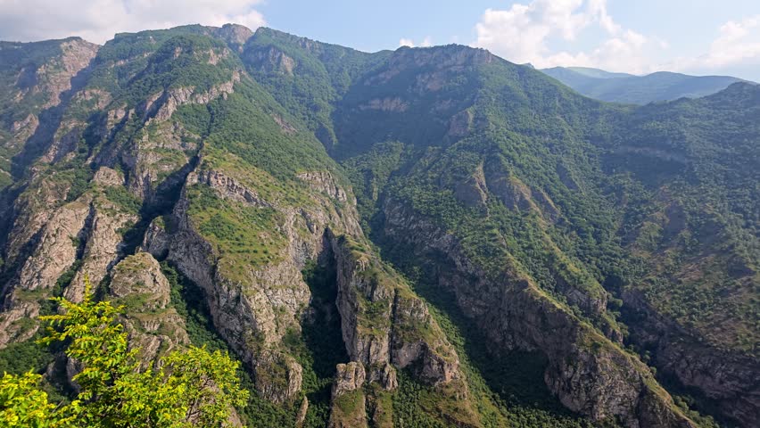 Green hills rise under blue sky with scattered clouds. Rocky peaks stand tall among dense vegetation