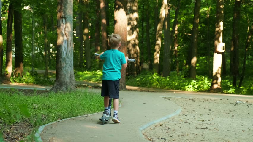 Kid riding a scooter in a forest park. Child riding a scooter outside in slow motion. Cute boy doing healthy sport activities outdoors. 
