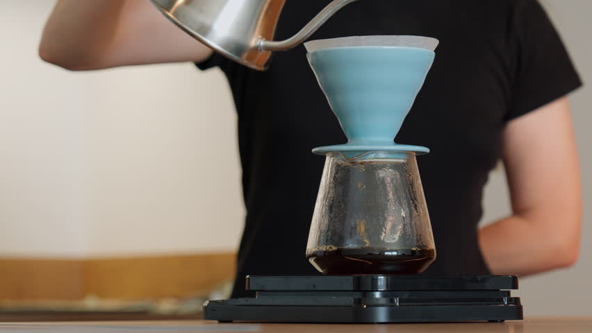 A woman pours hot water from a gooseneck kettle into a blue dripper with a paper filter. Fresh brew drips into a glass server on a kitchen counter.