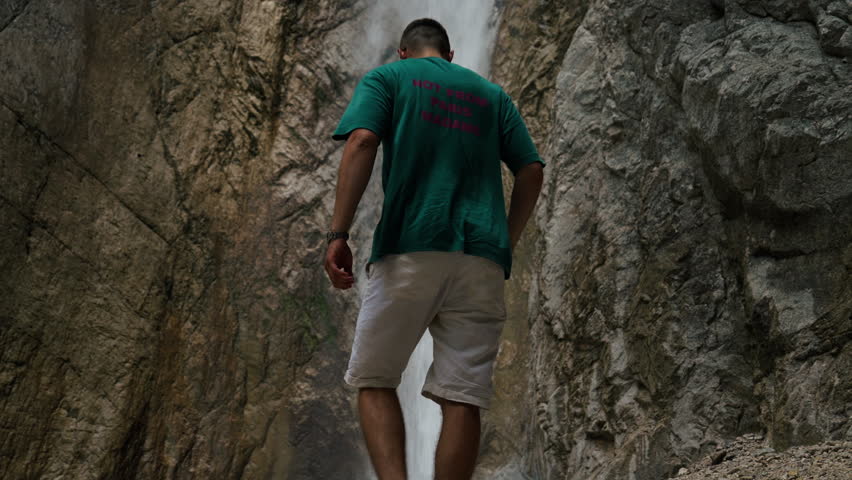 A young man stands close to a waterfall inside a narrow rocky gorge. Water drops behind him between steep stone walls.