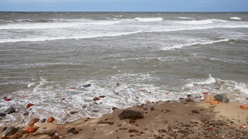 Waves gently washing over sandy beach scattered with various rocks and pebbles along rough sea shore. Overcast sky casts muted tones over dynamic coastal scene emphasizing natural seaside environment