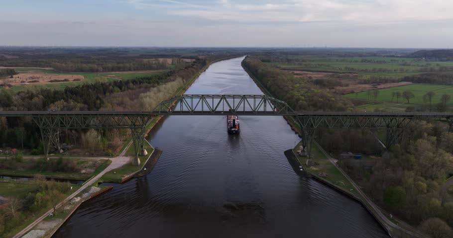 A drone view of large cargo ship passes under a metal railroad bridge spanning Kiel canal through lush green landscapes in Germany