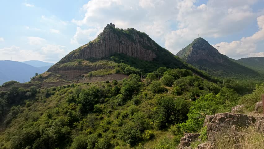 Green hills rise under blue sky with scattered clouds. Rocky peaks stand tall among dense vegetation