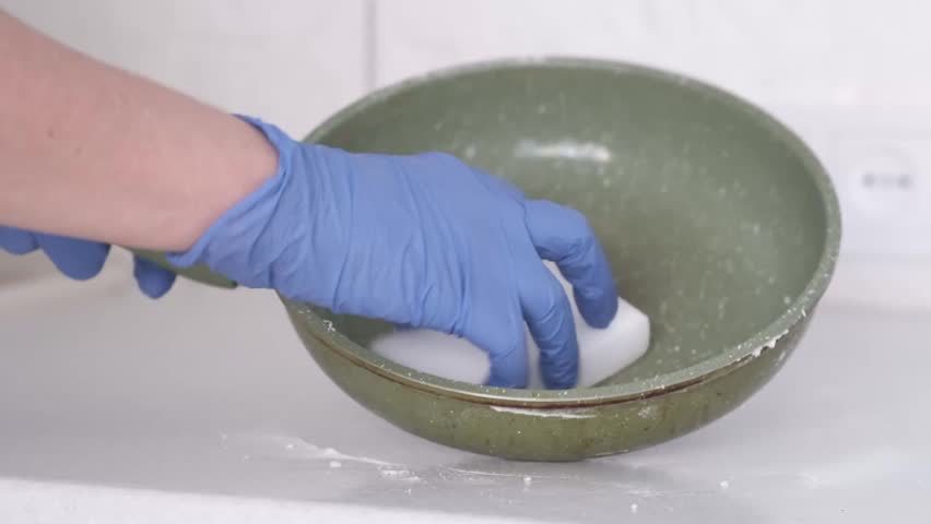 Close kitchen view shows blue gloved hand pressing white sponge inside green speckled frying pan, with stained inner surface, pale neutral background, soft daylight, practical home cleaning mood