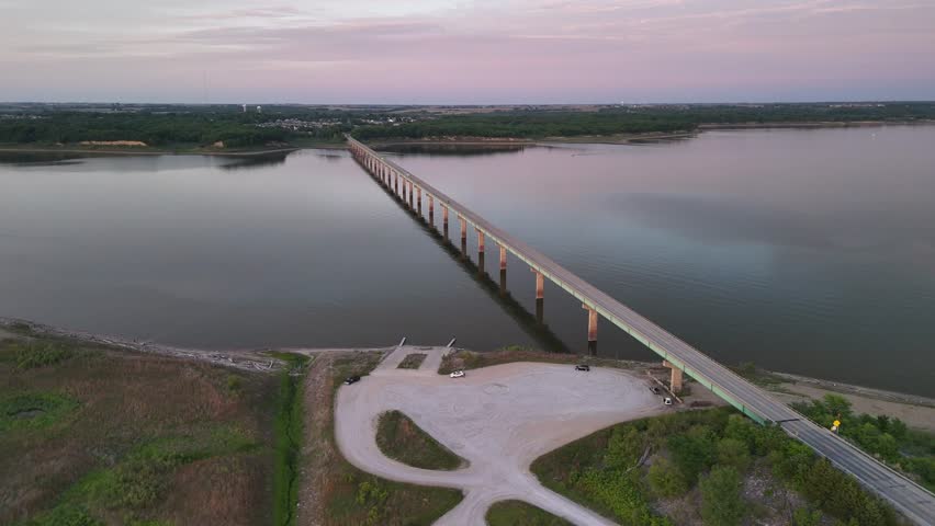 Cinematic aerial footage captures peaceful lake landscape featuring long bridge spanning across water surface during colorful sunset hour with clear sky and gravel parking lot below.