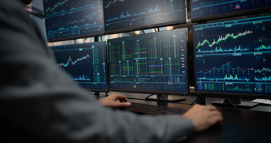 Over-the-shoulder View of a Male Trader Working at a Multi-monitor Desk Displaying Stock Exchange Data, Equity Charts and Market Analytics for Investment, Capital Growth. Asset Management Company