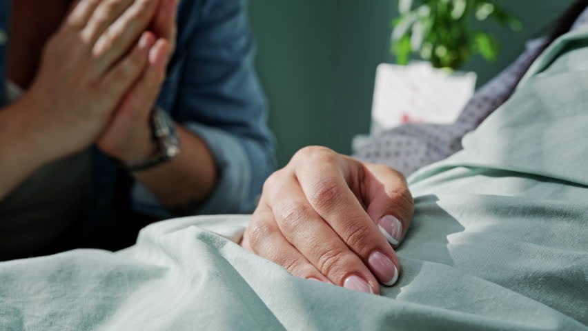 Man takes hand of beloved wife severe patient lying in bed in hospital closeup. Woman with dangerous disease rests and husband supports during treatment