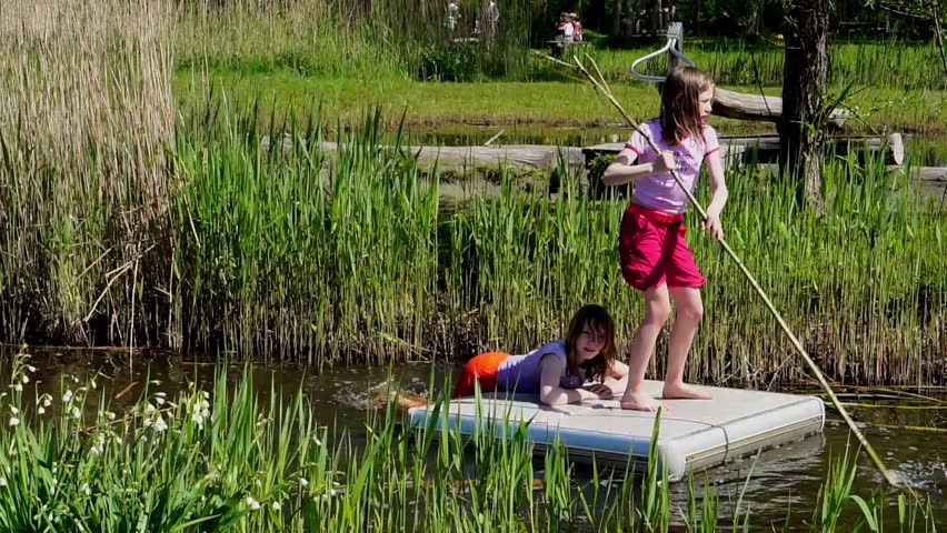 Two girls enjoy a playful rafting adventure on a small floating platform in a sunlit stream surrounded by reeds. One steers with a long pole while the other kicks her legs in the water, creating splashes. The moment captures summer freedom, friendship, and the joy of outdoor exploration.