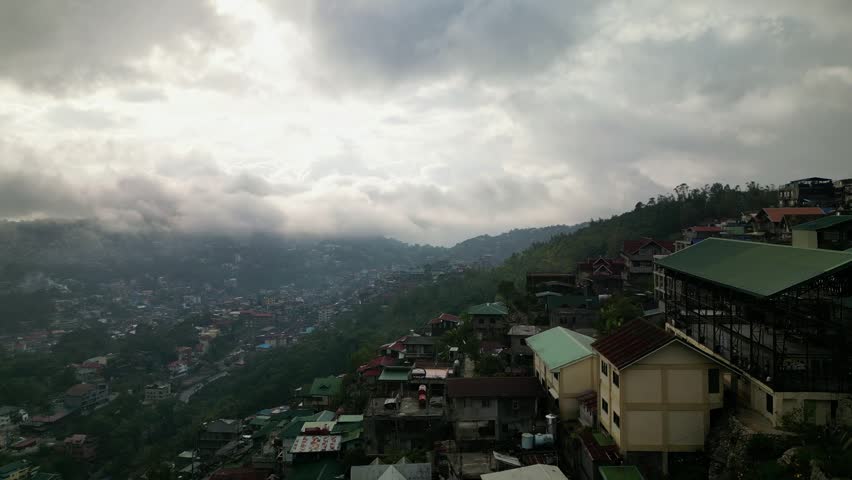 Dynamic aerial footage capturing sprawling urban landscape on a mountainside. Fog rolls in over distant cityscape under moody cloudy sky. Busy city life.
