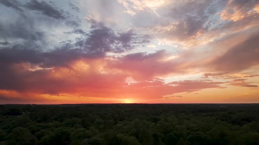 Wide-angle aerial view looking out across a lush green forest canopy towards a vibrant, colorful sunset with textured clouds in rural Oklahoma