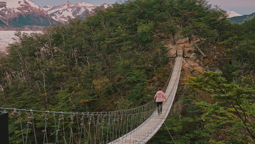 A woman walking across a suspended bridge surrounded by the natural beauty of forests, glaciers, and mountains in the distance, Torres del Paine, Patagonia, Chile 