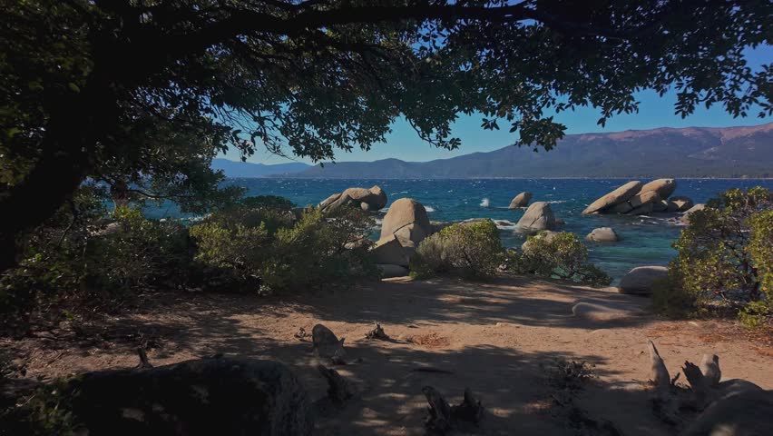Rocky shoreline at Lake Tahoe with blue water and mountains in California, framed by tree branches. Scenic natural landscape with waves and clear sky, ideal for travel and outdoor use